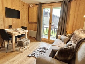 A cozy room with a wooden interior, a sofa, a dining table with chairs, and a view through a glass door.