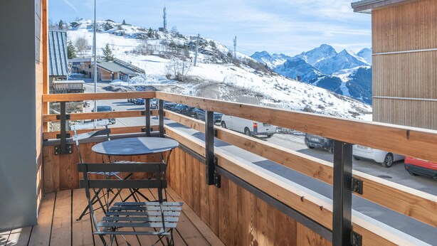 Une terrasse en bois avec une table et des chaises, offrant une vue sur un paysage montagneux enneigé et un espace de stationnement.