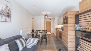 Un salon moderne avec un canapé gris, une table à manger avec des chaises, une cuisine avec des meubles en bois et une bibliothèque remplie de livres et d’objets décoratifs.
