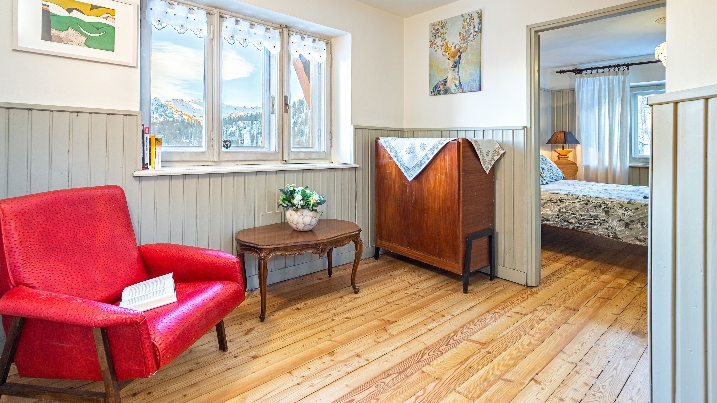 A cozy room with a red armchair, a small table with a vase of flowers, a wooden cabinet, and a bed with a patterned bedspread.