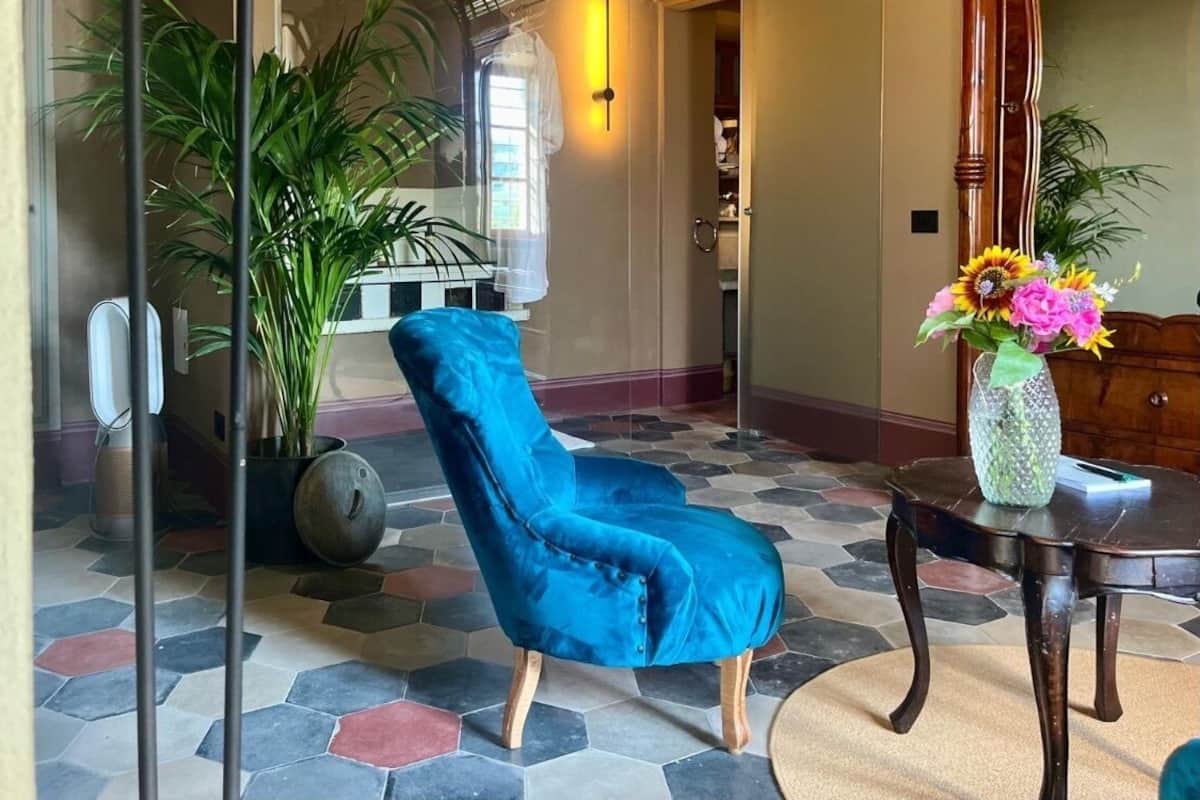 Una habitación con una butaca de terciopelo azul, una planta en maceta, un jarrón con flores y una mesa de madera.
