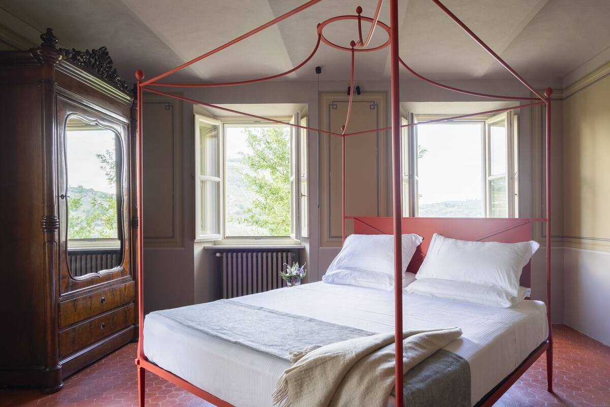 Una cama con dosel y cuatro columnas, ropa de cama blanca, un aparador de madera y vista a follaje exterior.