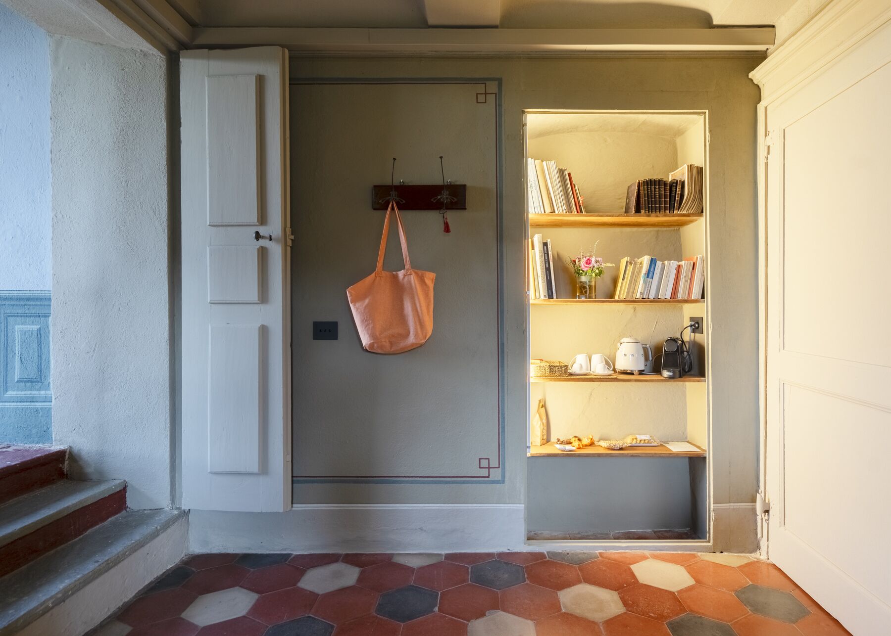 A hallway with a door leading to a room containing a bookshelf, a small table with cups and a kettle, and a bag hanging on the wall.