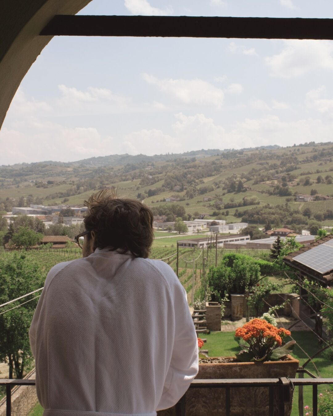 A person standing on a balcony overlooking a rural landscape with rolling hills and scattered buildings.