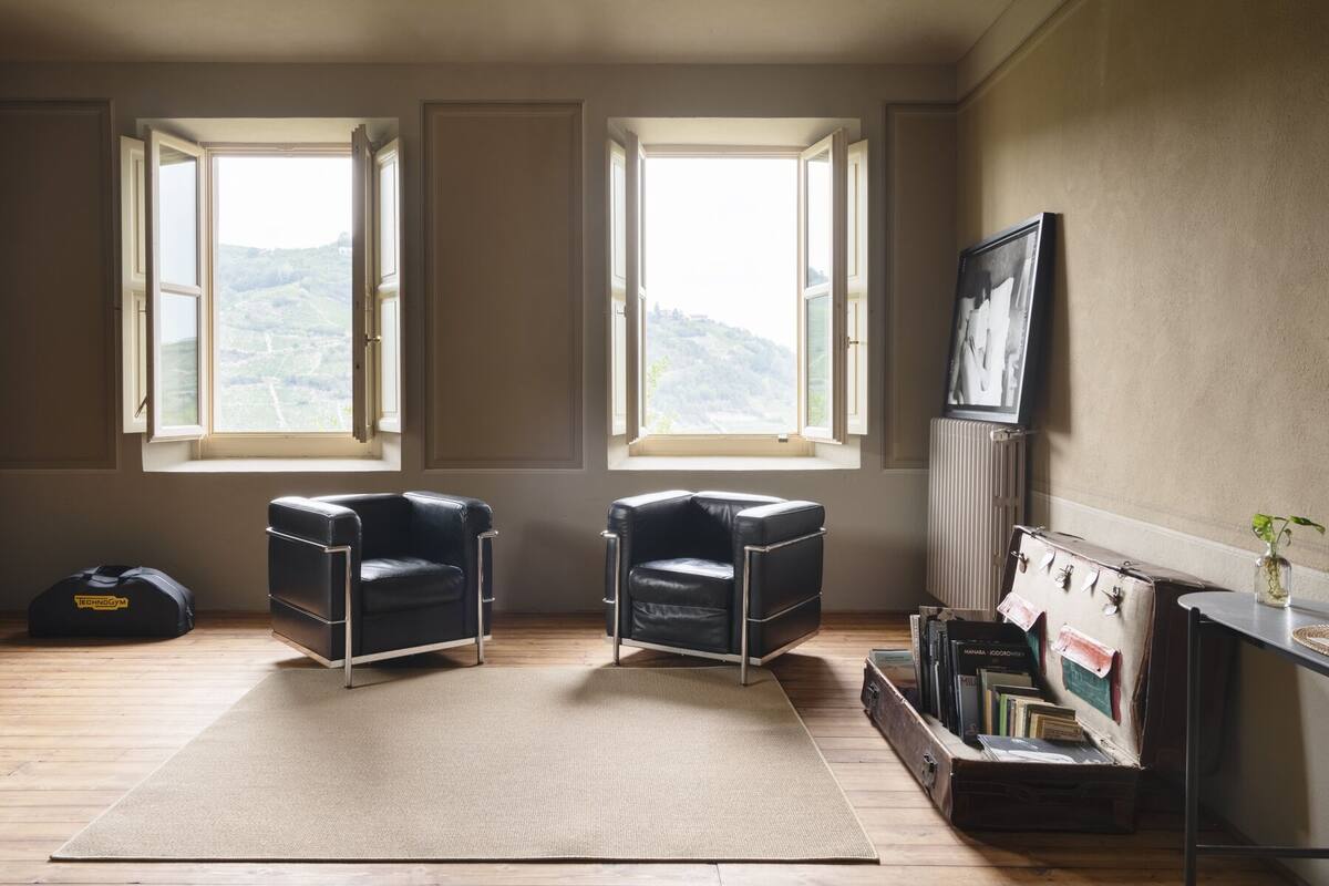 Una habitación con dos sillones de cuero negro, un ventanal con vistas a colinas, un radiador y una estantería con libros y un cuadro enmarcado.