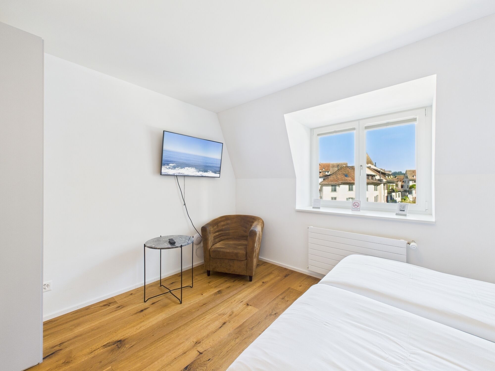 Une chambre à coucher comprenant un lit, une chaise, une petite table, un téléviseur fixé au mur et une fenêtre donnant sur des immeubles.
