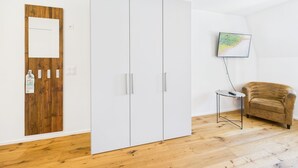 Ein Zimmer mit Holztür, einem weißen Kleiderschrank, einem kleinen Tisch mit Fernbedienung, einem braunen Sessel und einem an der Wand montierten Fernseher.