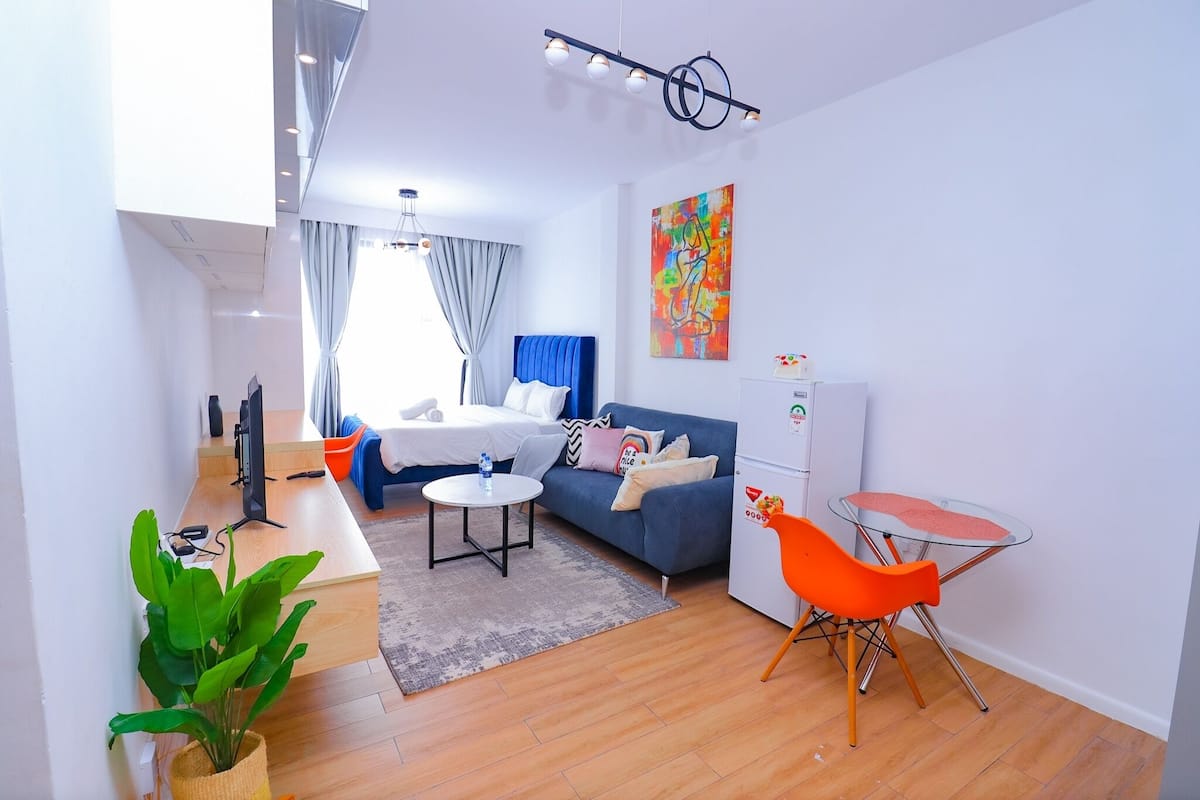 Une pièce moderne et bien éclairée, avec un canapé bleu, une petite table ronde et un fauteuil orange vif. Une grande fenêtre, ornée de rideaux, laisse entrer la lumière naturelle, et un tableau abstrait aux couleurs vives décore le mur.