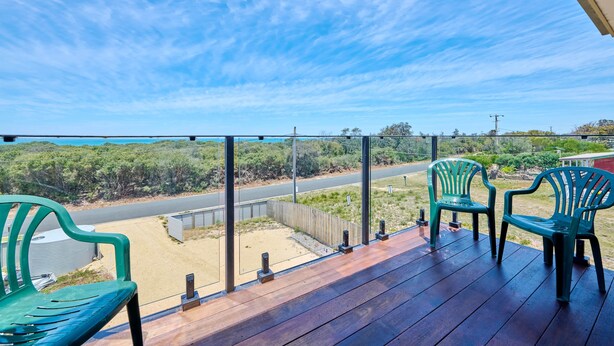 Una terraza de madera con sillas verdes, vista a una carretera y vegetación al fondo.