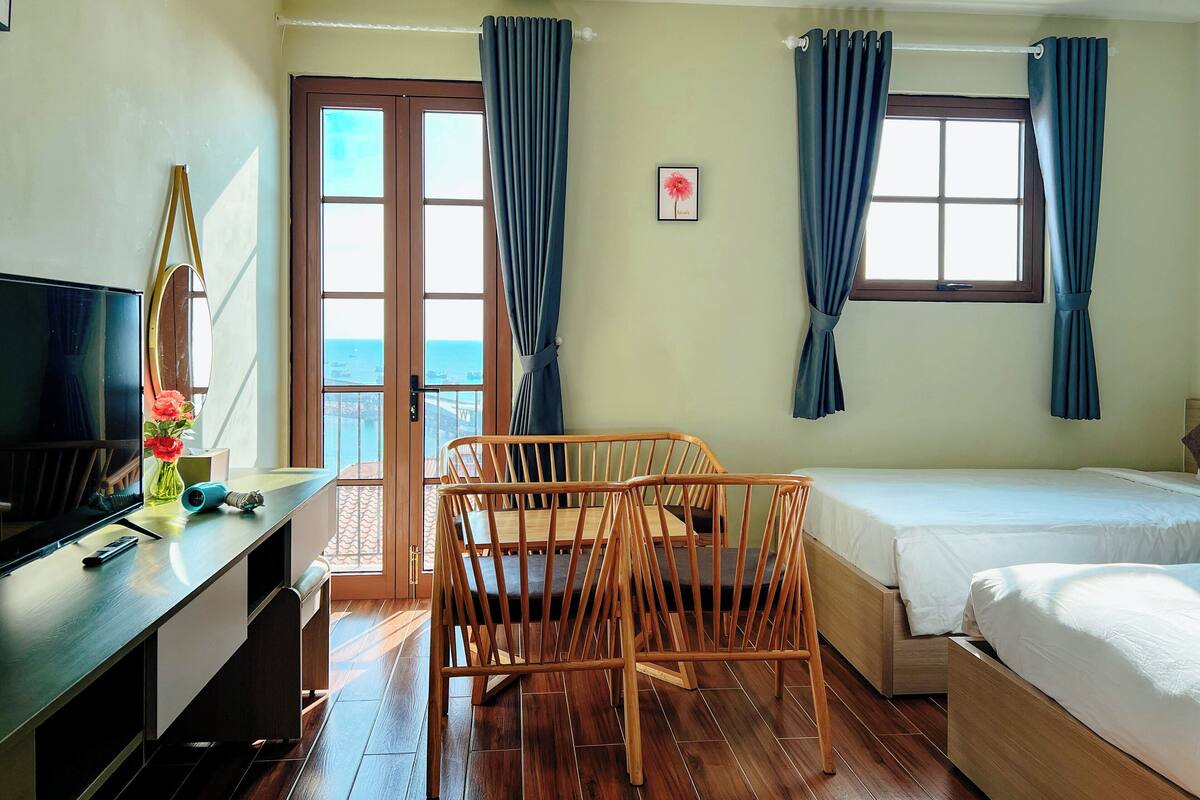 A hotel room with a bed, a desk with a TV, a chair, and a view of the sea through a door.