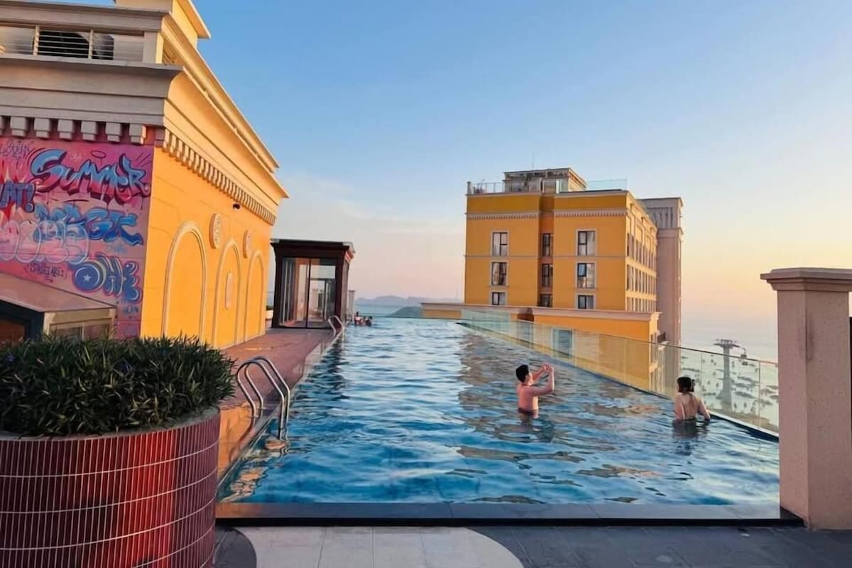 An infinity pool with a view of a cityscape, a covered seating area, and a building with a yellow facade.