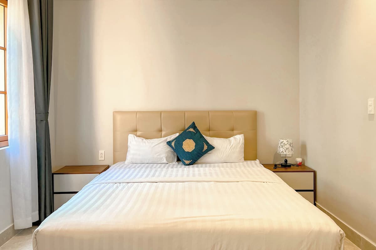 A neatly made bed with white bedding and a beige headboard. There are two bedside tables, one with a lamp and a small decorative item.