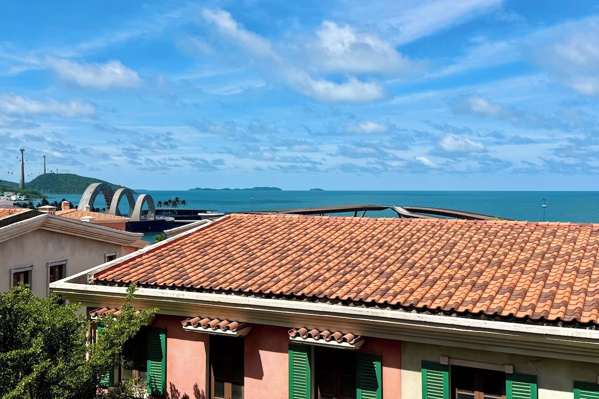 A coastal building with a tiled roof, green shutters, and a view of the sea and a bridge in the background.