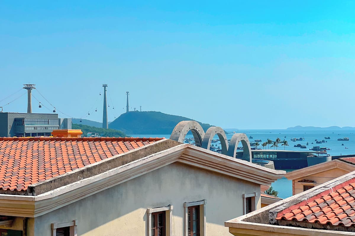 A coastal cityscape with a prominent cable car system, a large arch structure, and a view of the sea with boats.