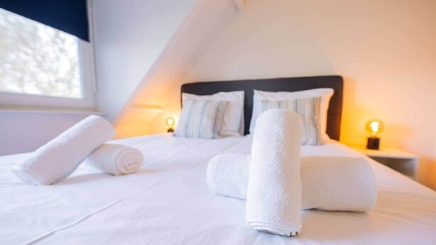 Cama bem arrumada com toalhas brancas dobradas, cabeceira escura e luminárias laterais.