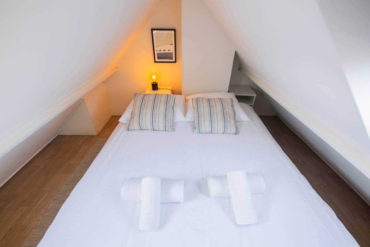 A neatly made bed with pillows and rolled towels in a small, sloped room with a single bedside lamp and a framed picture on the wall.