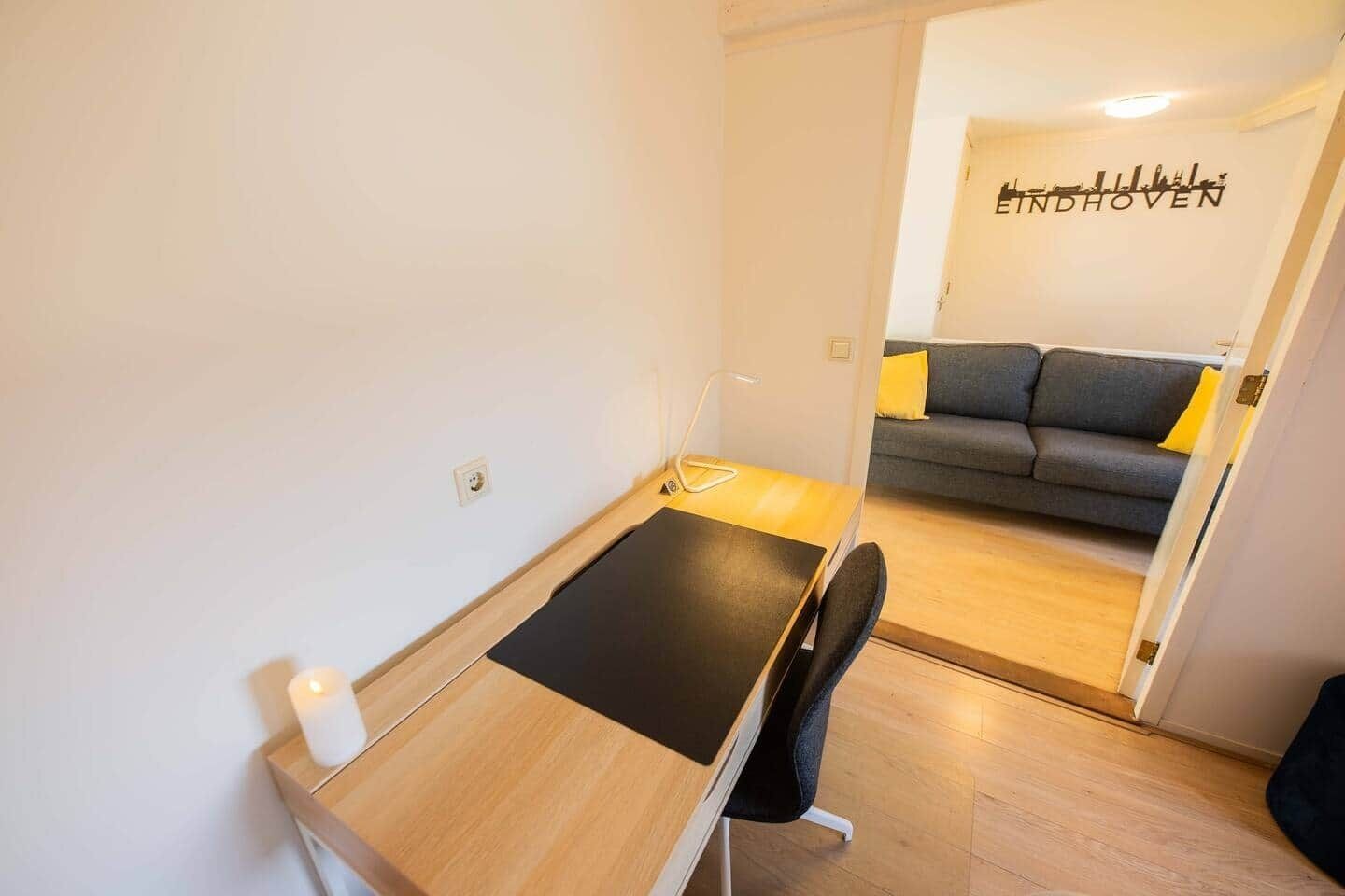 A compact room with a wooden desk, a chair, and a small table. A sofa with yellow cushions is visible in the background.