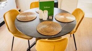 Mesa redonda com quatro jogos americanos trançados, duas cadeiras amarelas e um letreiro de boas-vindas verde.
