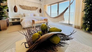 A bedroom with a hammock, a bed with white linens, a large window with a view, and a fruit bowl on the floor.