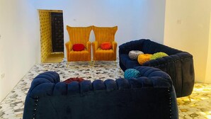 A room with a patterned floor, a dark blue upholstered sofa, two yellow armchairs with red cushions, and a small ottoman. - Flenjo Zone Bar and Apartments (Ado Ekiti)