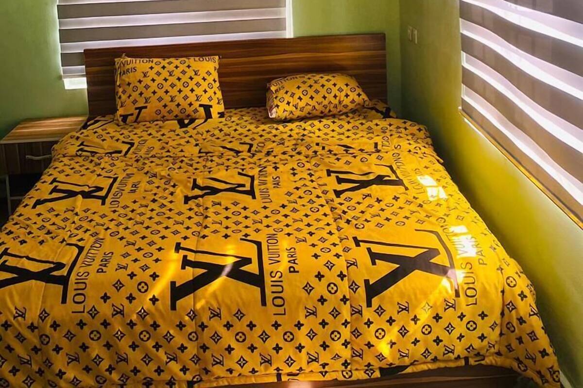Une chambre à coucher avec un lit recouvert d’une literie jaune ornée d’un motif Louis Vuitton, deux oreillers, une tête de lit en bois et une fenêtre avec des stores.