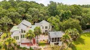 Une grande maison à deux étages, dotée d’une terrasse rouge, entourée d’une végétation luxuriante et d’arbres.