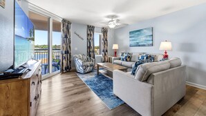 A living room with a sofa, coffee table, and a TV. There is a balcony with curtains and a view of the outdoors. - Spacious 2nd row condo, great amenities + Free Attraction Tickets! (North Myrtle Beach)