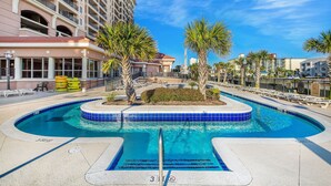 A hotel pool area with a clear blue pool, palm trees, and a large building in the background. - Spacious 2nd row condo, great amenities + Free Attraction Tickets! (North Myrtle Beach)