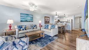 A living room with a sofa, coffee table, and lamps; a dining area with a table and chairs; a kitchen with cabinets and appliances; and a ceiling fan. - Spacious 2nd row condo, great amenities + Free Attraction Tickets! (North Myrtle Beach)