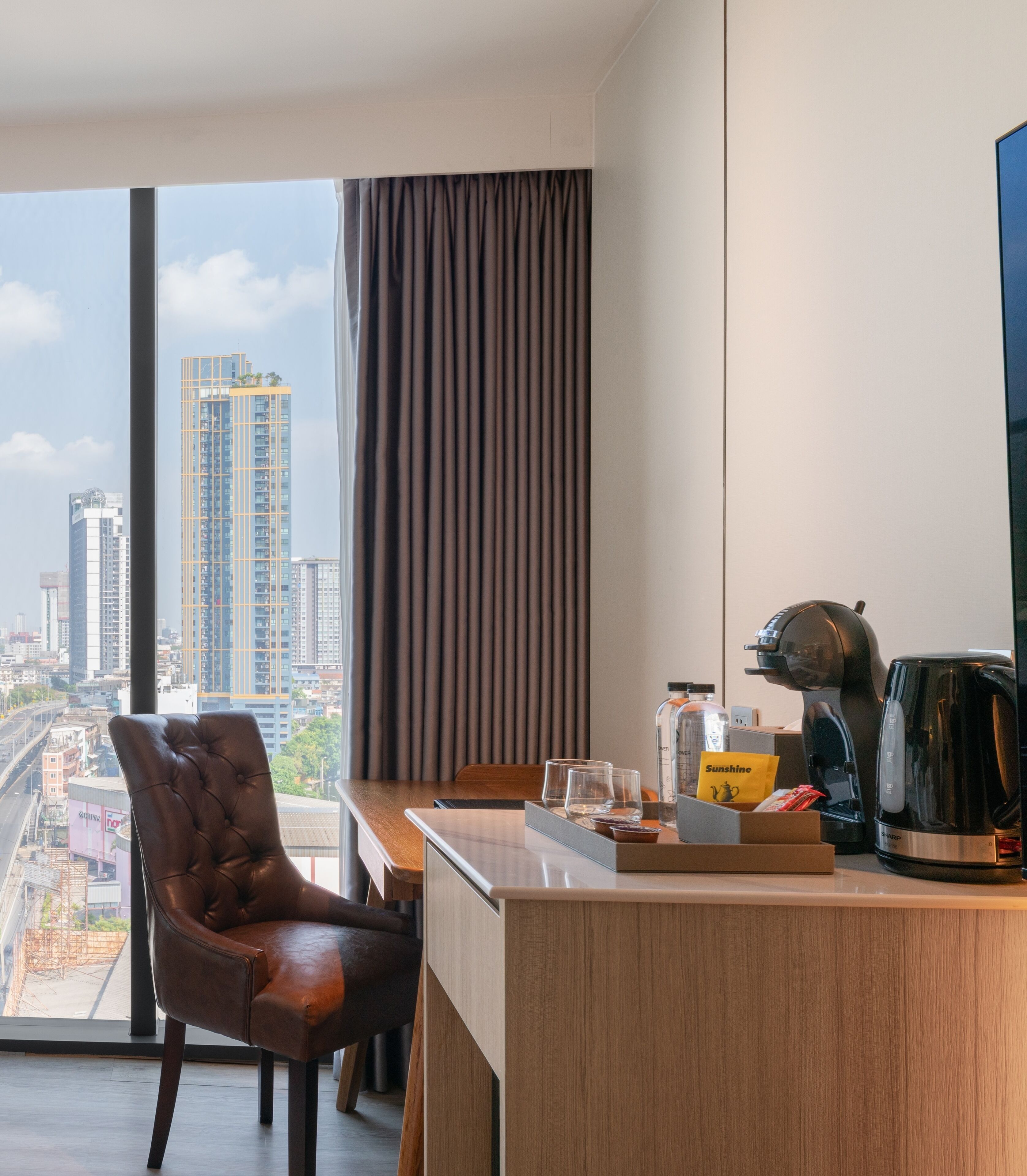 A modern hotel room with a city view, a leather chair, a wooden desk, and a coffee maker.