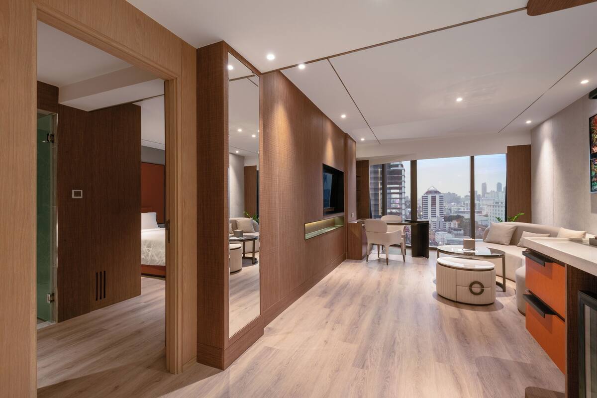A modern hotel room with a wooden floor, a sofa, a dining table, and a large window offering a city view.