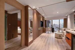 A modern hotel room with a wooden floor, a sofa, a dining table, and a large window offering a city view. - SZ CANALSIDE HOTEL (Bangkok)
