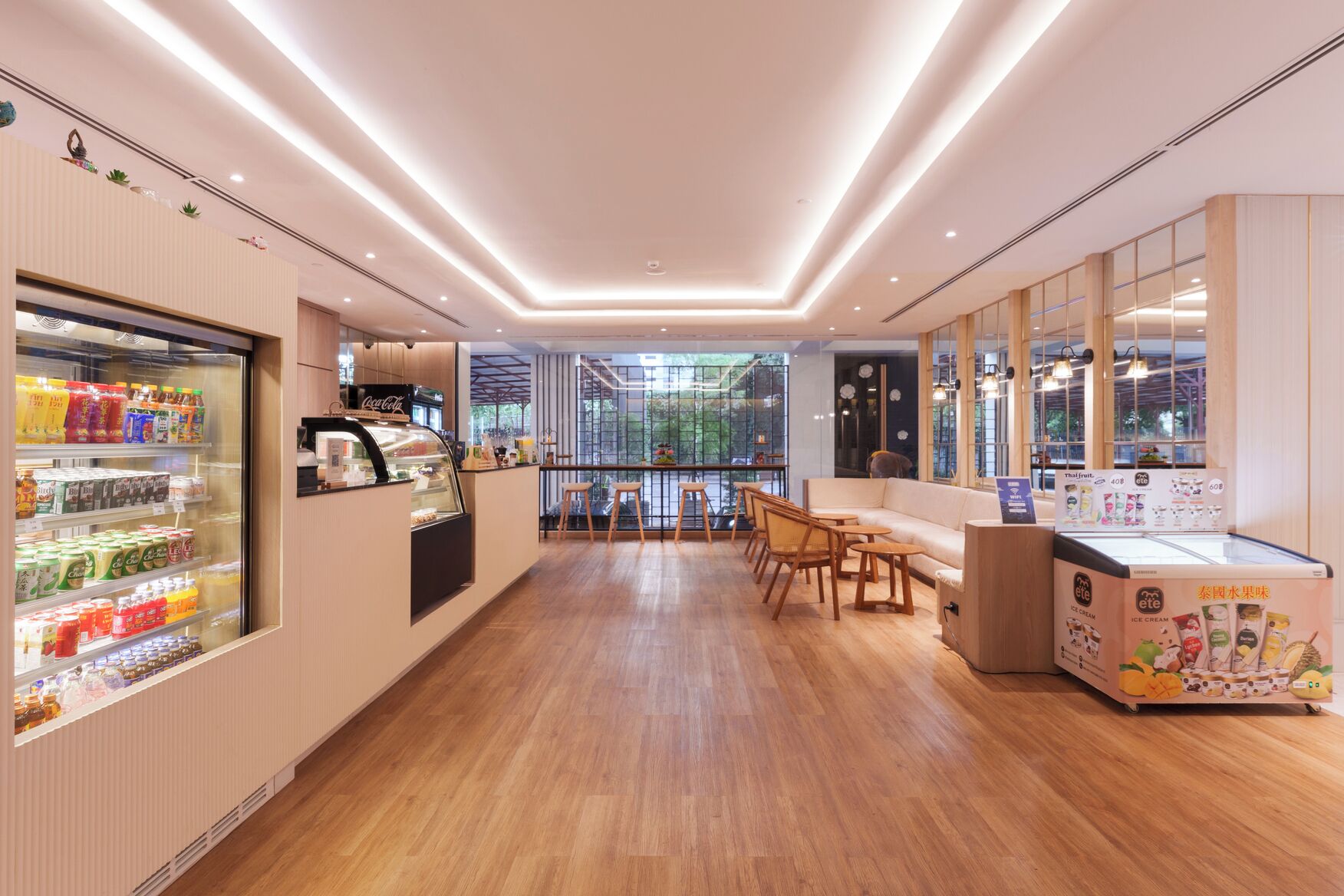 A modern cafe with a wooden floor, a seating area with tables and chairs, a refrigerated display case with beverages, and a large window offering a view of the outdoors.