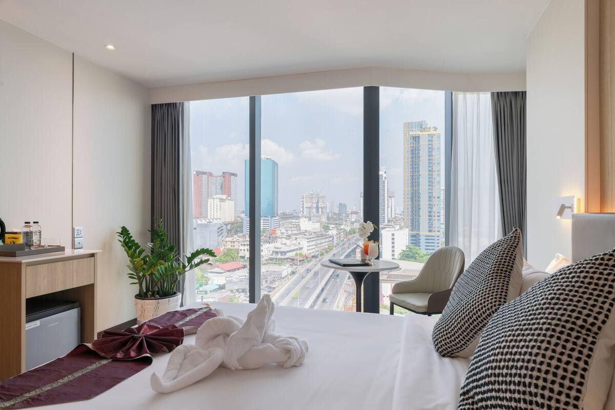 A hotel room with a large bed, a desk, a chair, a small table, and a potted plant.