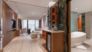 Een moderne hotelkamer met een groot bad, een eethoek met een ronde tafel en stoelen, een kleine koelkast, en uitzicht op de stad.