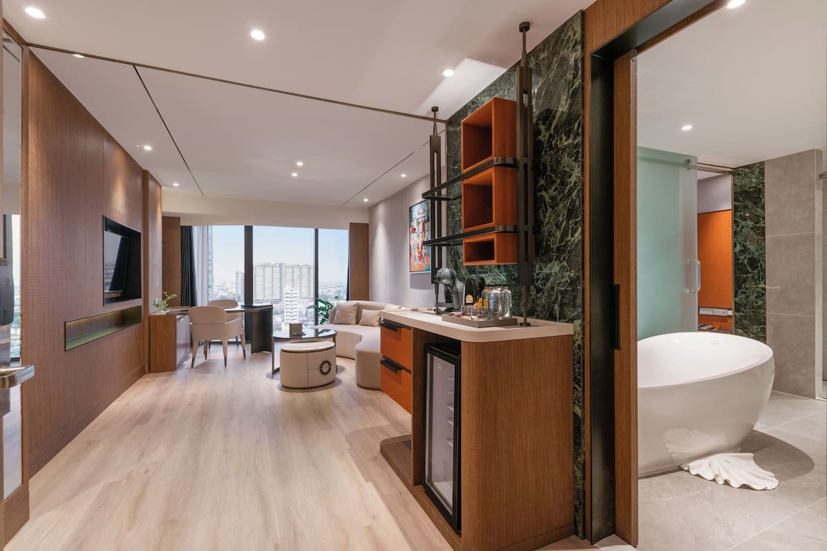 A modern hotel room with a large bathtub, a dining area with a round table and chairs, a small refrigerator, and a view of the city.