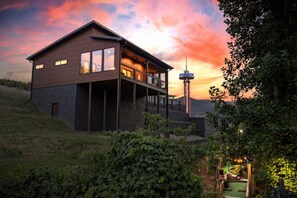 Una casa moderna de dos pisos con balcón, rodeada de follaje y un cielo al atardecer.
