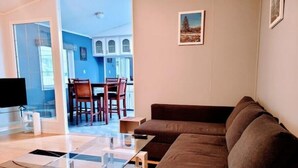 Ein Wohnzimmer mit einem braunen Sofa, einem Glastisch und einem Fernseher auf einem Schrank. Durch eine offene Tür ist ein Essbereich zu sehen.