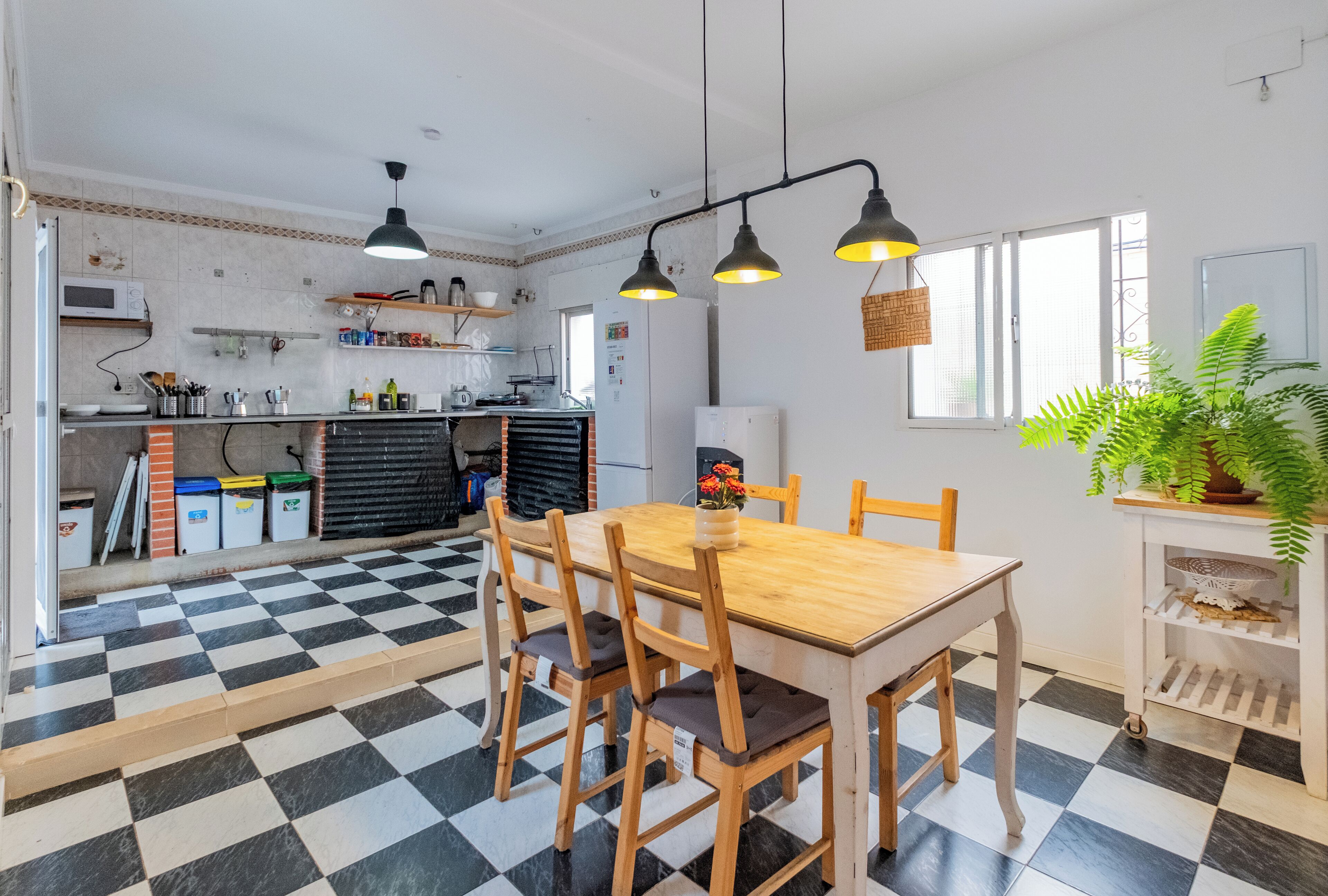 Eine Küche mit Schachbrettboden, einem Holzespeisetisch und Stühlen. Es befinden sich Hängeleuchten, ein Kühlschrank und eine Mikrowelle darin.