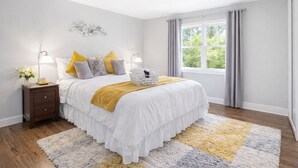 Een slaapkamer met een bed, nachtkastje, lamp en uitzicht op groen buiten.