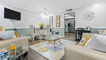 A modern living room with a glass coffee table, beige sofas, and a dining area with a chandelier.
