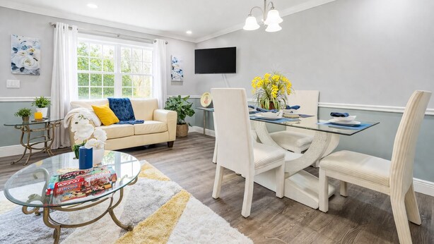 Een moderne woonkamer met een glazen salontafel, een bank met kussens, een eettafel met stoelen, en een flatscreen televisie die aan de muur is bevestigd.