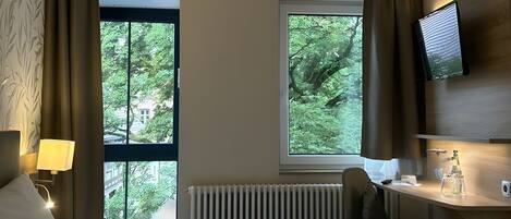 A hotel room with a bed, a desk, a chair, a TV, and a window with a view of trees.