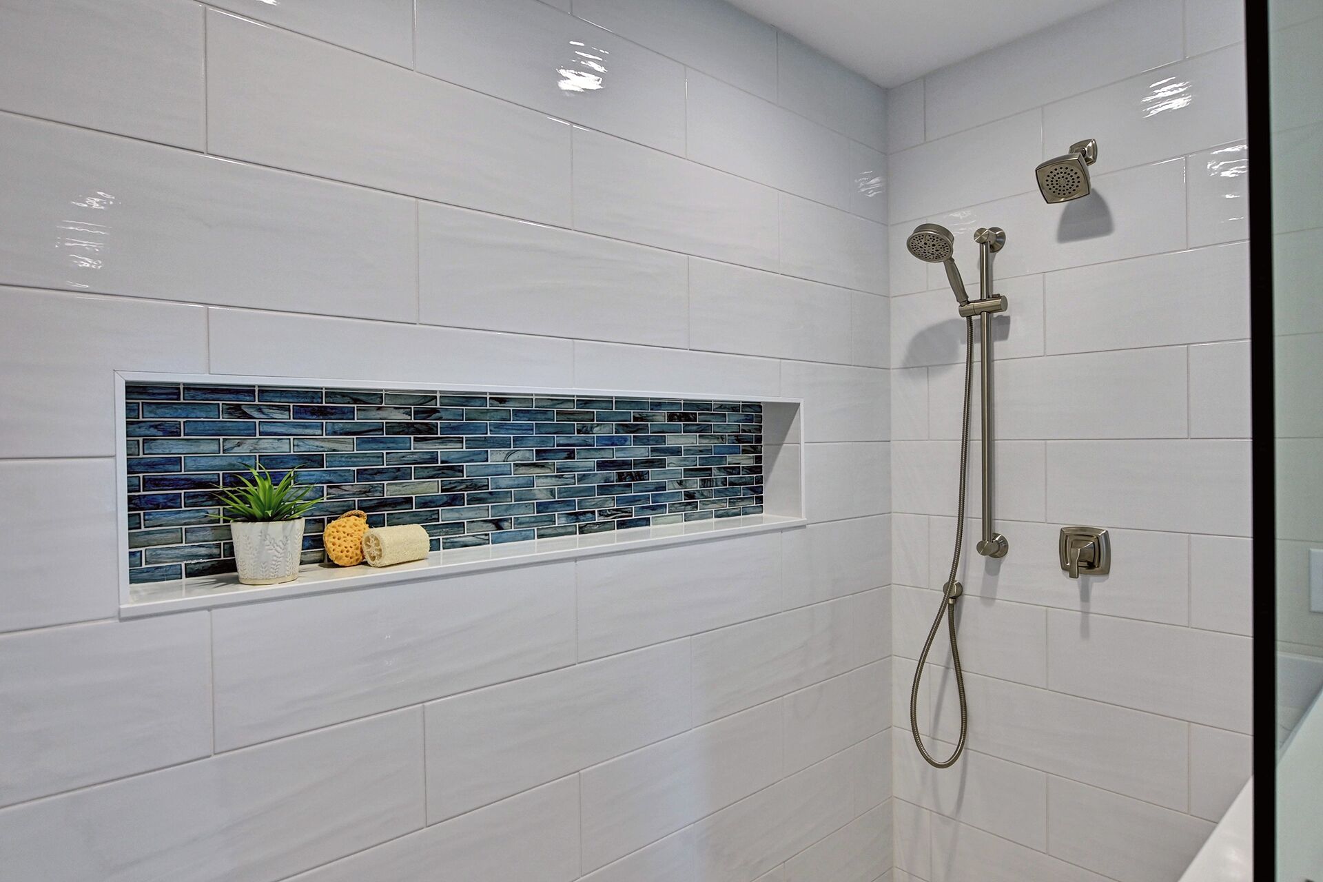 Un baño con azulejos blancos, un estante azulado y una ducha con cabezal de ducha manual, además de un panel de control instalado en la pared.