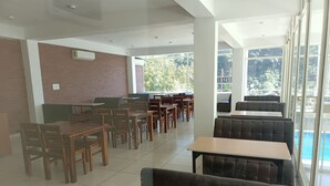 Un espace intérieur vaste et lumineux, avec des tables et des chaises en bois, un mur en briques et de grandes fenêtres donnant sur l’extérieur.