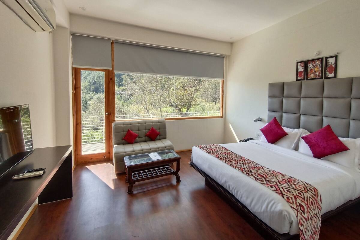 A bedroom with a bed, a sofa, a small table, and a window with a view of trees.