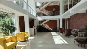 Un hall d’entrée spacieux avec un canapé jaune, un fauteuil marron et un ensemble de meubles pour le salon. On y trouve un escalier avec un mur en briques rouges, et une personne se tient sur la plateforme supérieure.
