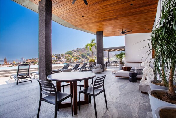 Un patio al aire libre con una mesa redonda y sillas, una zona de estar con sofás y una estatua, y vista a la playa y edificios al fondo.