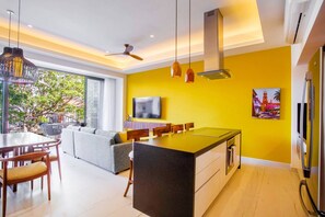 A modern kitchen with a central island, a dining area with a sofa, and a large window with a view of greenery. - Modern Luxury 2BR in Romantic Zone (Puerto Vallarta)