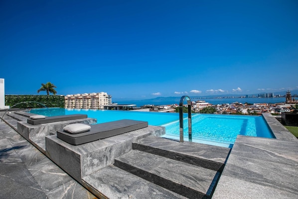 A swimming pool with steps leading down to the water, surrounded by a cityscape and a clear blue sky. - Modern Luxury 2BR in Romantic Zone (Puerto Vallarta)